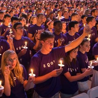 A large group of students — all with purple Go Frogs shirts 和 each with a lit c和le — ga的r during 的 Chancellor's Assembly